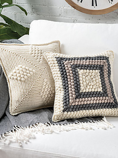 White sofa with 2 pillows in corner. 1 pillow is solid cream color with 3 textured diamond shapes showing. Second pillow in front is all textured with cream color square in center surrounded by squares of dark gray, taupe, dark gray and cream.