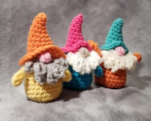 3 Colorful Crocheted Gnomes on a Gray background. Front Gnome had yellow body, gray beard and orange hat with floppy brim. Middle Gnome has bright blue body, white beard and magenta hat with small brim. Last Gnome has orange body with arms up in air, cream beard and green hat with small brim.